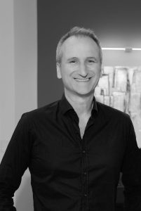 Ritratto professionale in bianco e nero di Michele Volpi Interior Designer sorridente con camicia scura, davanti a uno sfondo sfocato di uno studio di design.