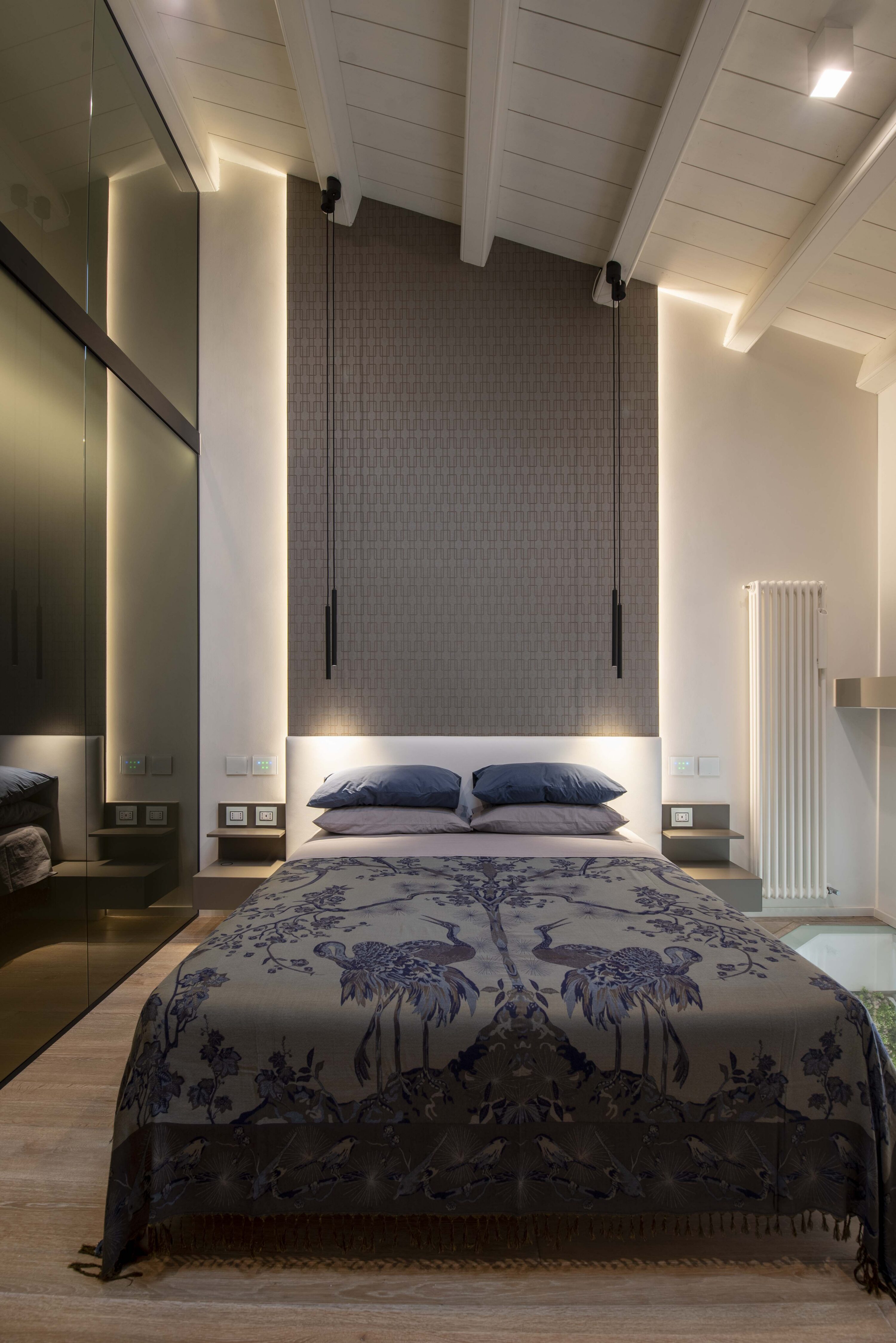 Vista frontale simmetrica di un letto matrimoniale con copriletto jacquard a motivi di gru, testiera retroilluminata a LED, parete d'accento tessile, lampade a sospensione tubolari e armadio in vetro sulla sinistra.