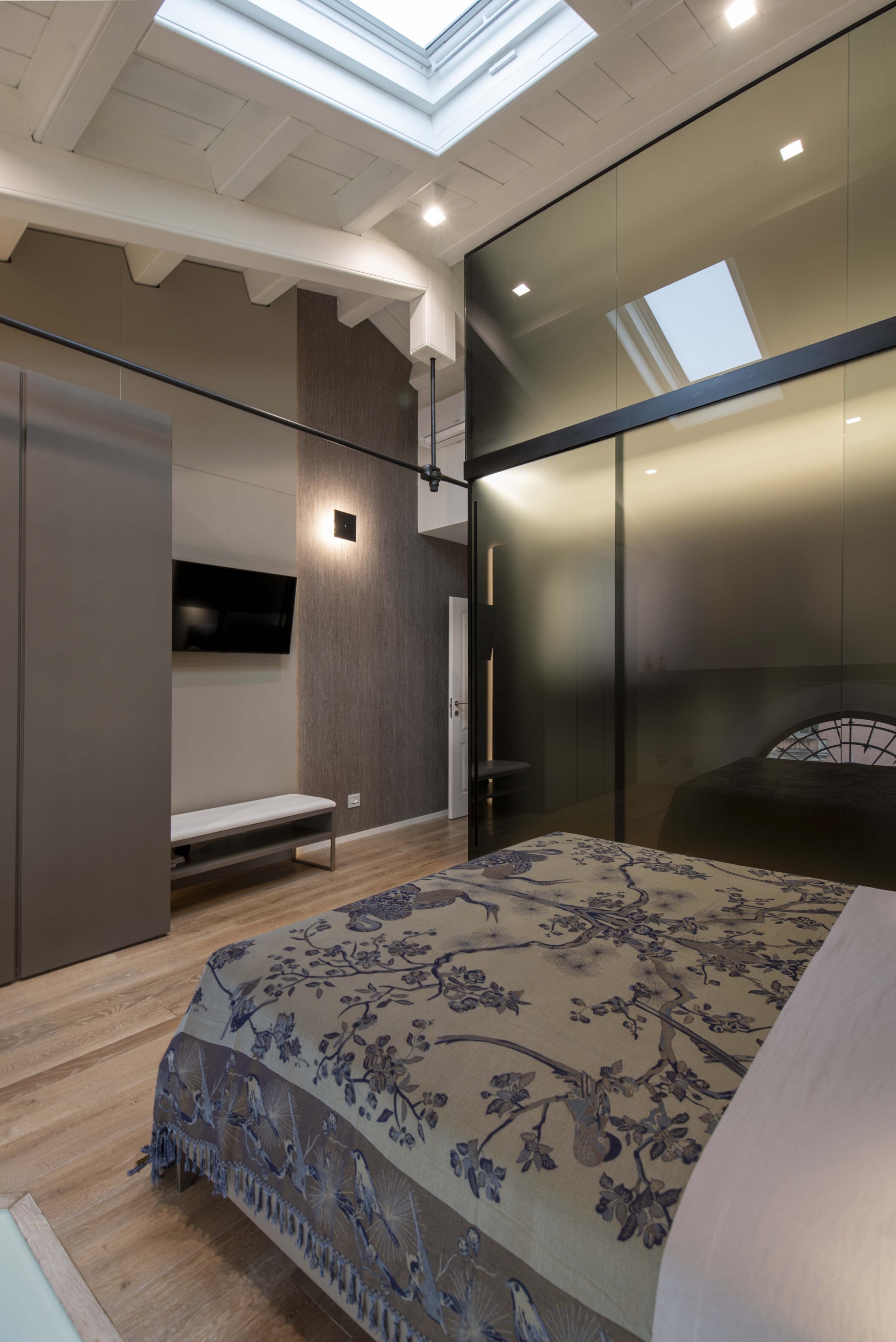 Prospettiva della camera da letto padronale con letto matrimoniale in primo piano, armadio a parete con ante in vetro fum&eacute;, TV a muro e soffitto con travi bianche e lucernario.