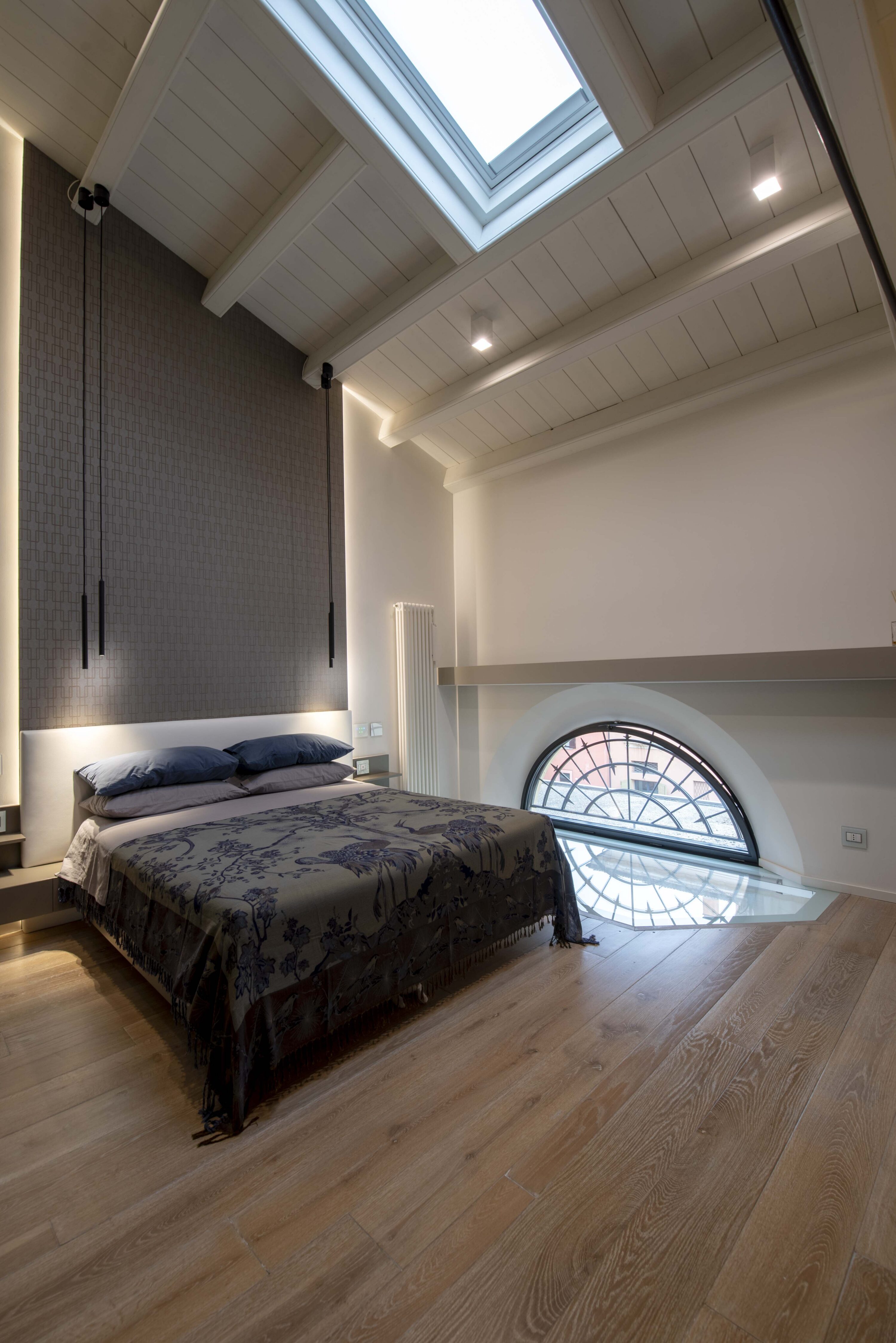 Camera matrimoniale soppalcata con letto al centro, finestra a lunetta ad arco sul pavimento in vetro, lucernario sul soffitto a travi bianche e parete di fondo decorata.