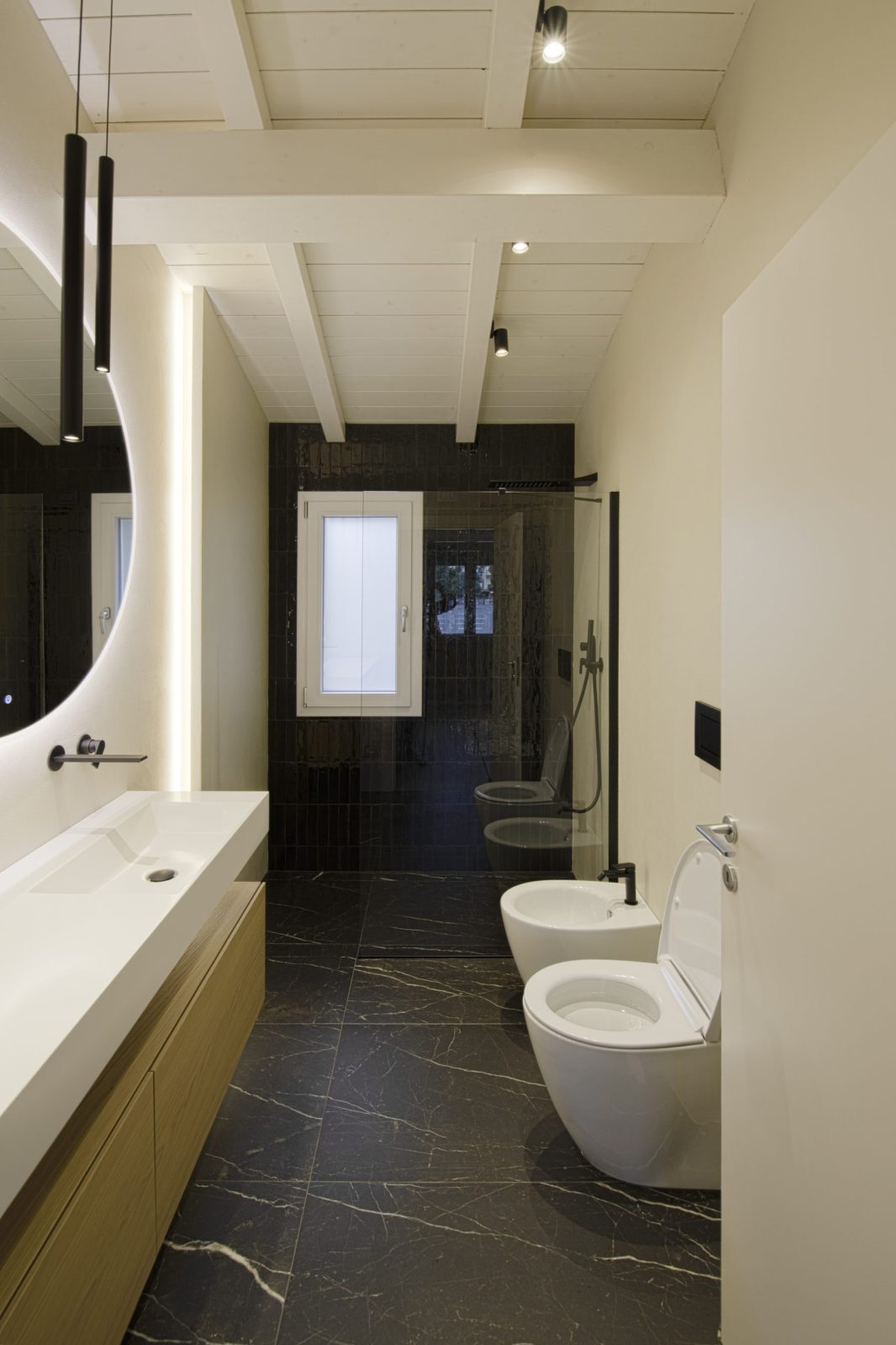 Bagno moderno con pavimento e doccia in gres nero venato, mobile lavabo in legno, grande specchio tondo retroilluminato e soffitto a travi bianche.
