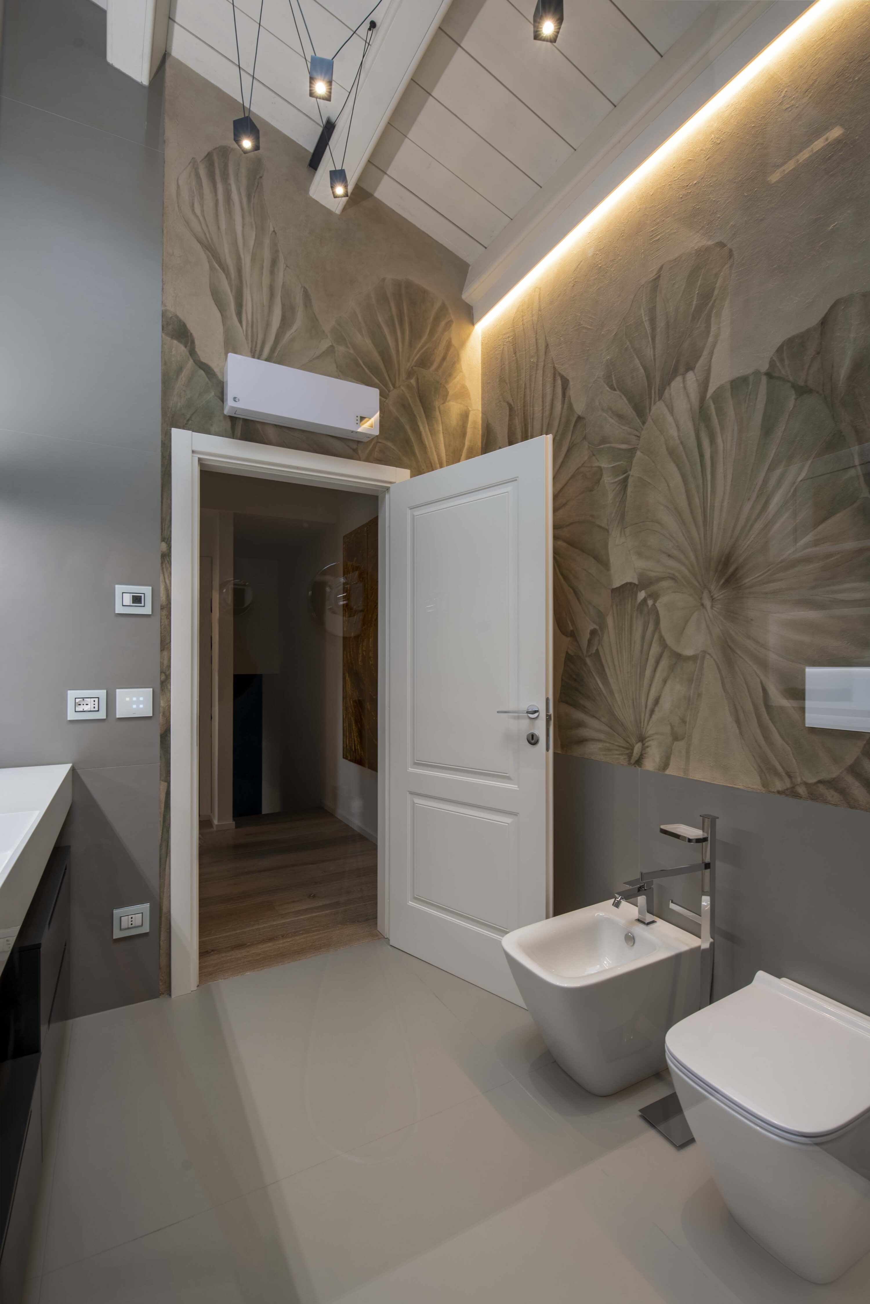 Bagno padronale moderno con sanitari sospesi bianchi, parete con carta da parati a grandi foglie, luce a LED e soffitto con travi in legno bianco.