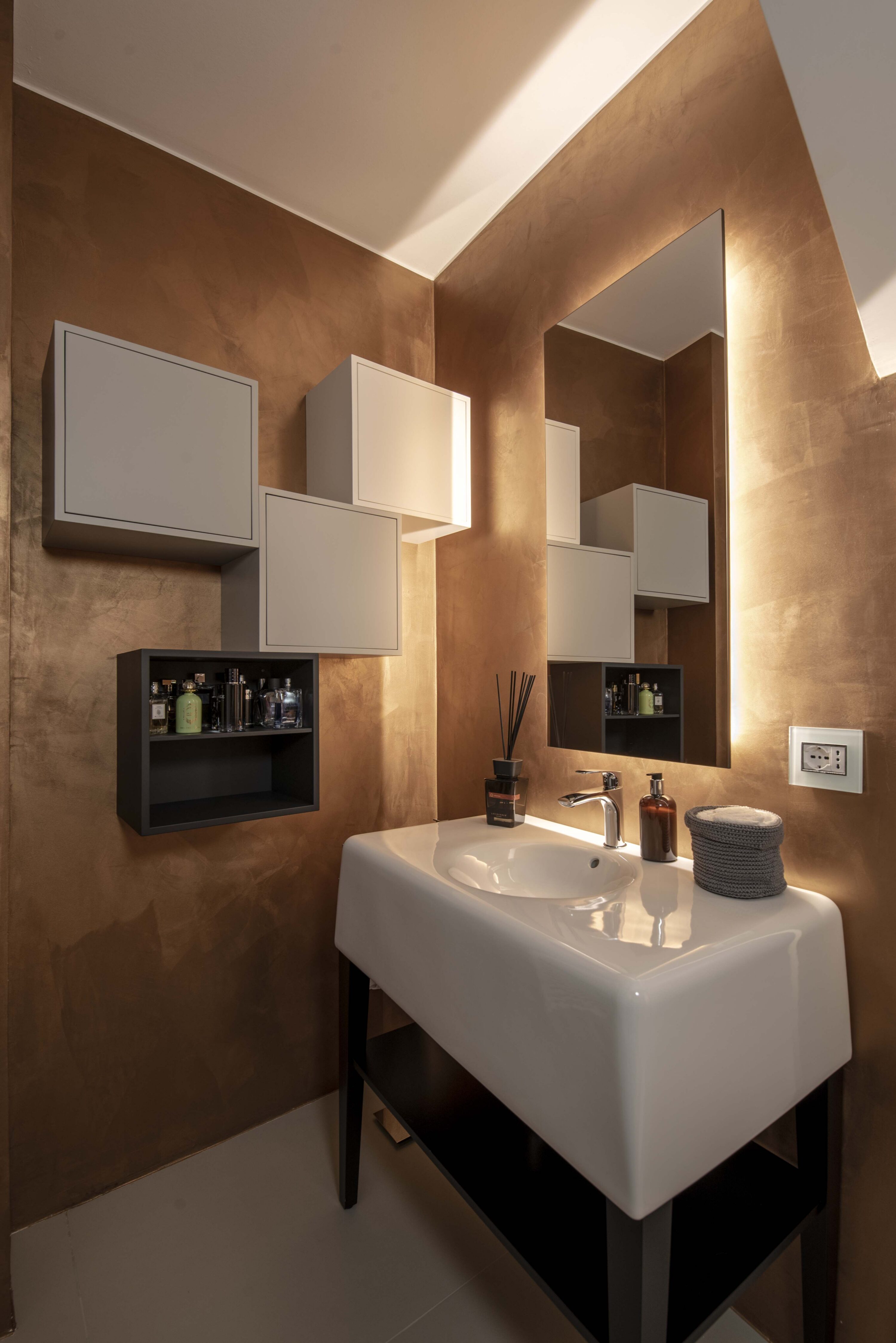 Angolo bagno design con lavabo freestanding bianco su struttura nera, pareti in resina bronzo lucido, specchio retroilluminato e pensili sospesi asimmetrici.