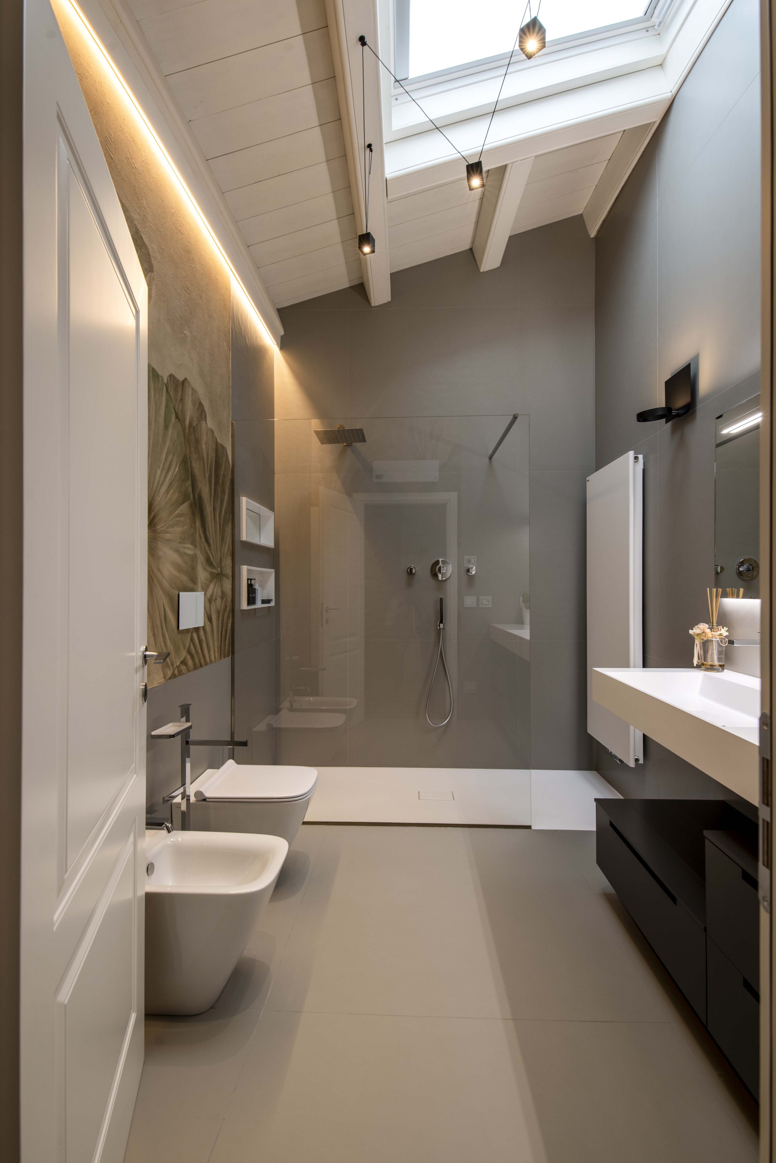 Prospettiva completa di un bagno padronale moderno in mansarda con ampia doccia walk-in in fondo, lucernario, sanitari sospesi, carta da parati botanica e lavabo su mobile scuro.