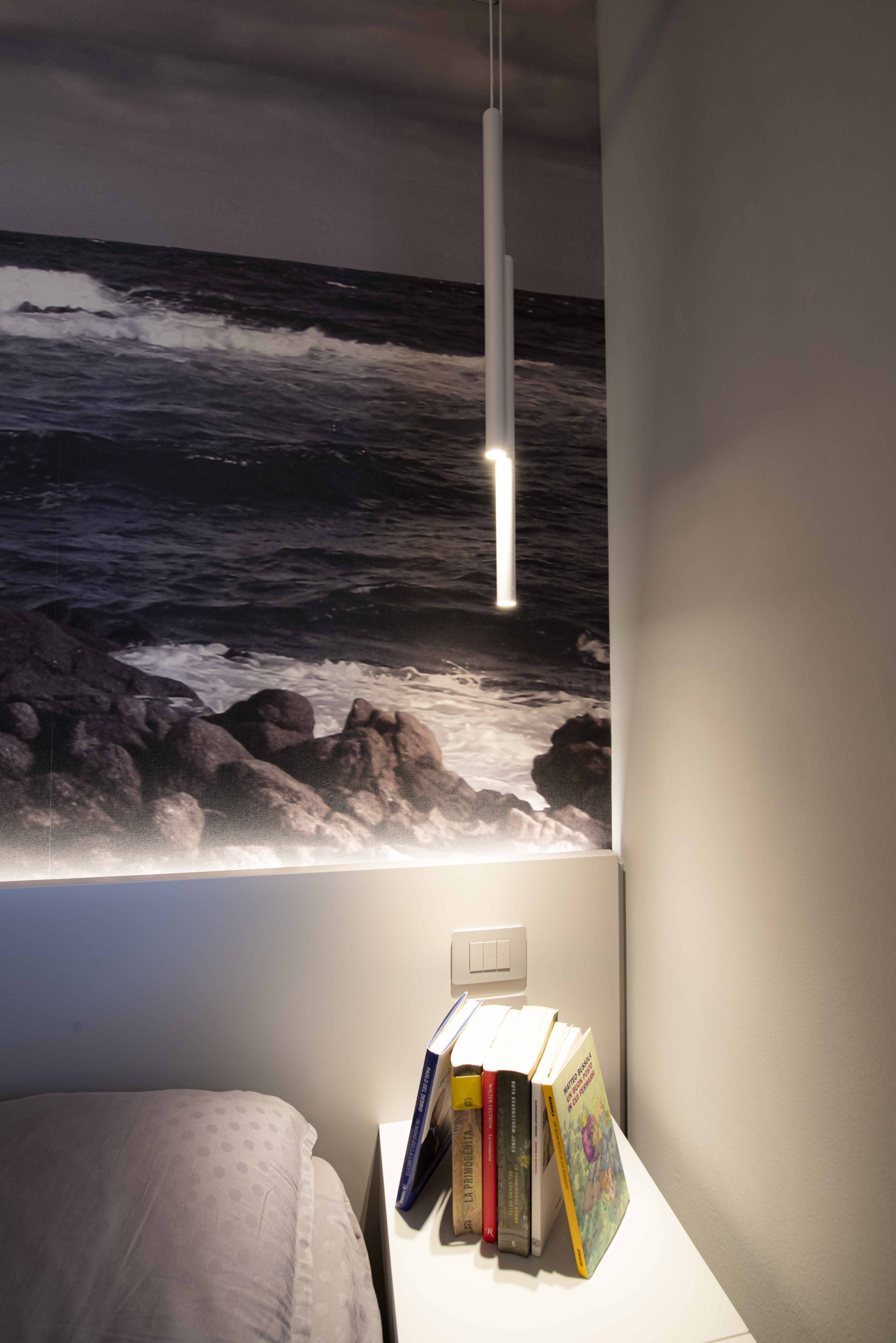 Primo piano di un comodino bianco con una selezione di libri per gli ospiti e lampada LED a sospensione sopra il letto.
