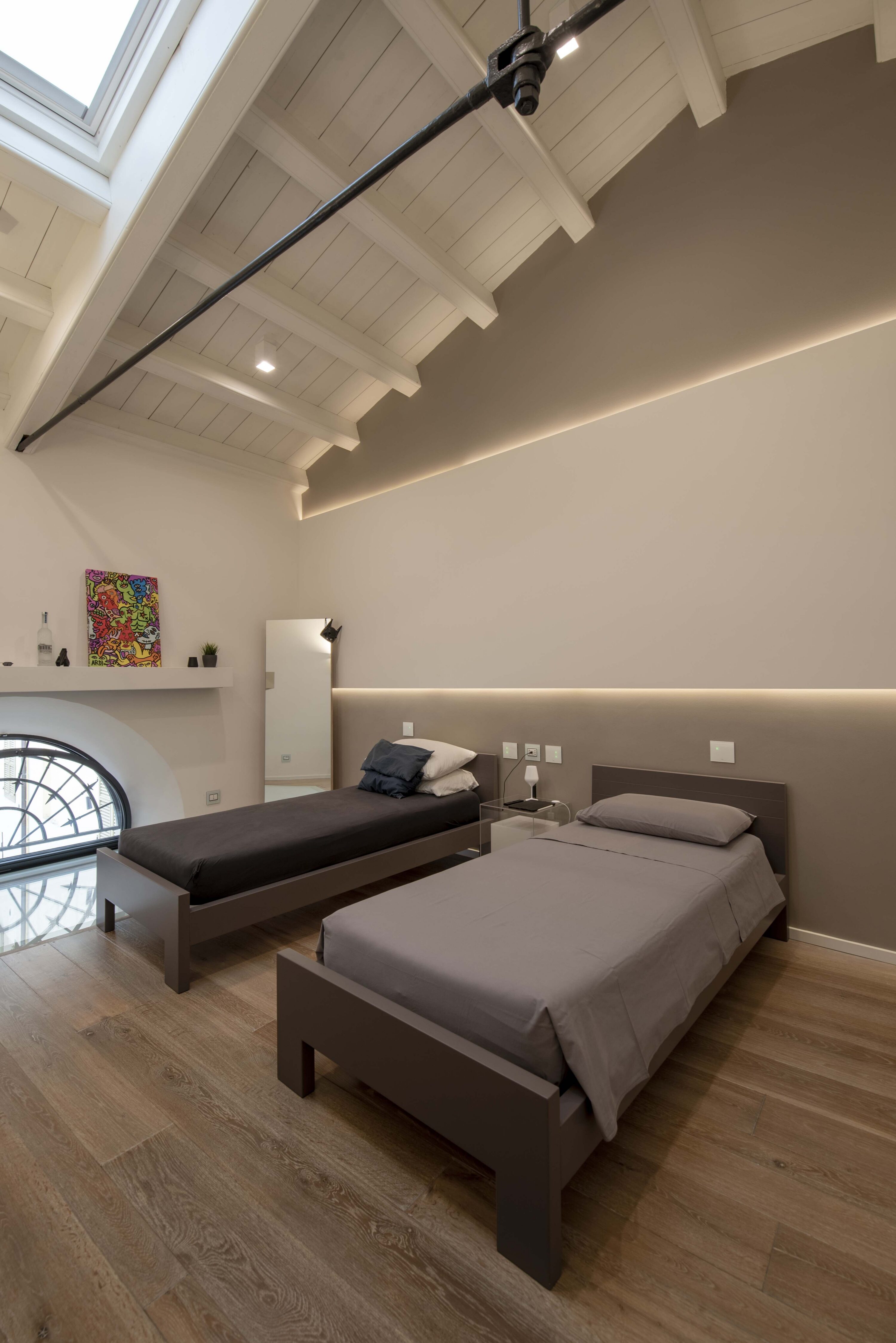 Camera da letto in mansarda moderna con due letti singoli grigi e neri, pavimento in legno, soffitto con travi bianche e lucernario, e luce LED a incasso lungo la parete.