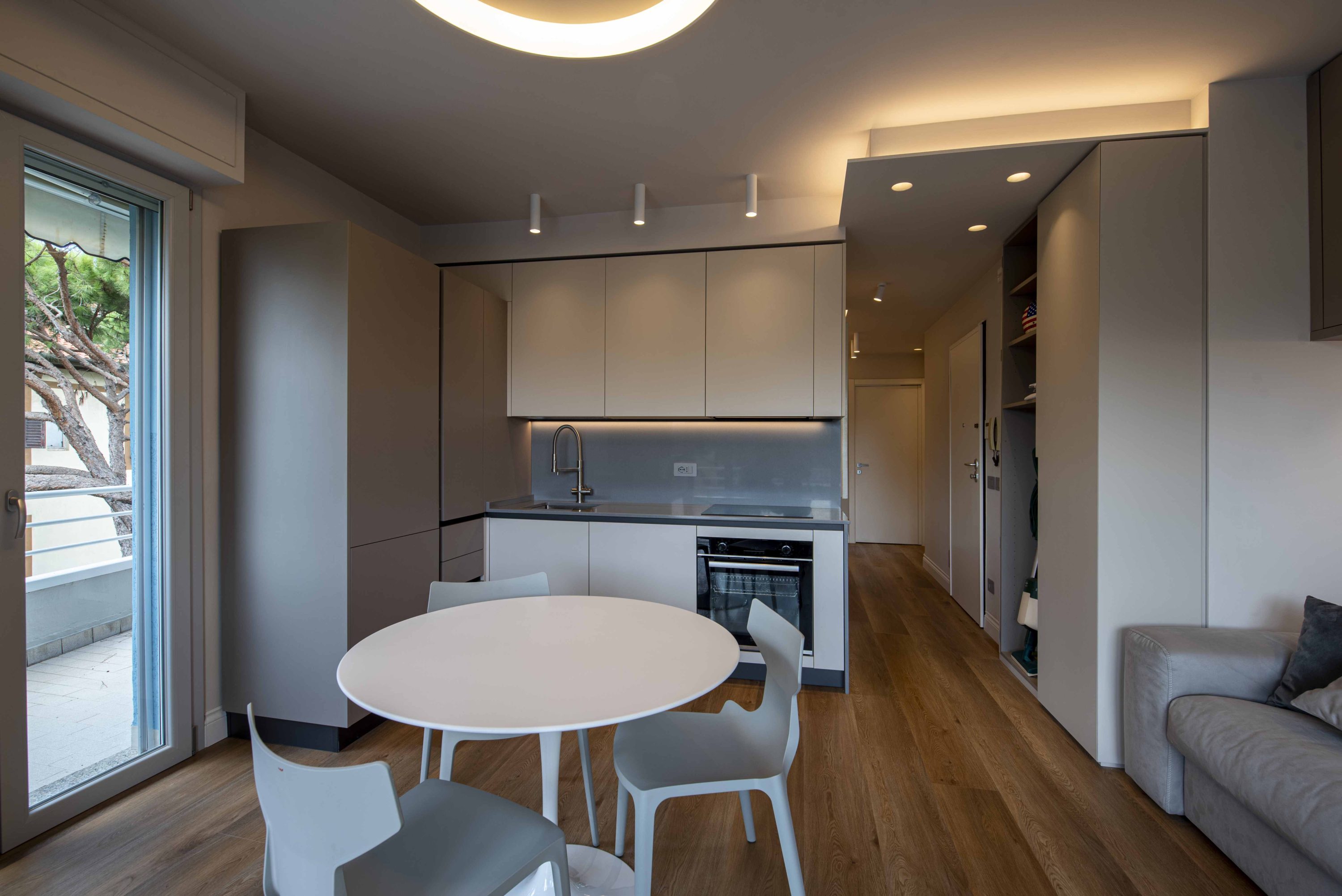 cucina-soggiorno-open-space-moderno-parquet – MICHELE VOLPI STUDIO INTERIOR DESIGN Open space moderno con cucina lineare bianca e grigia, tavolo tondo, sedie di design, pavimento in parquet e vista sul balcone.