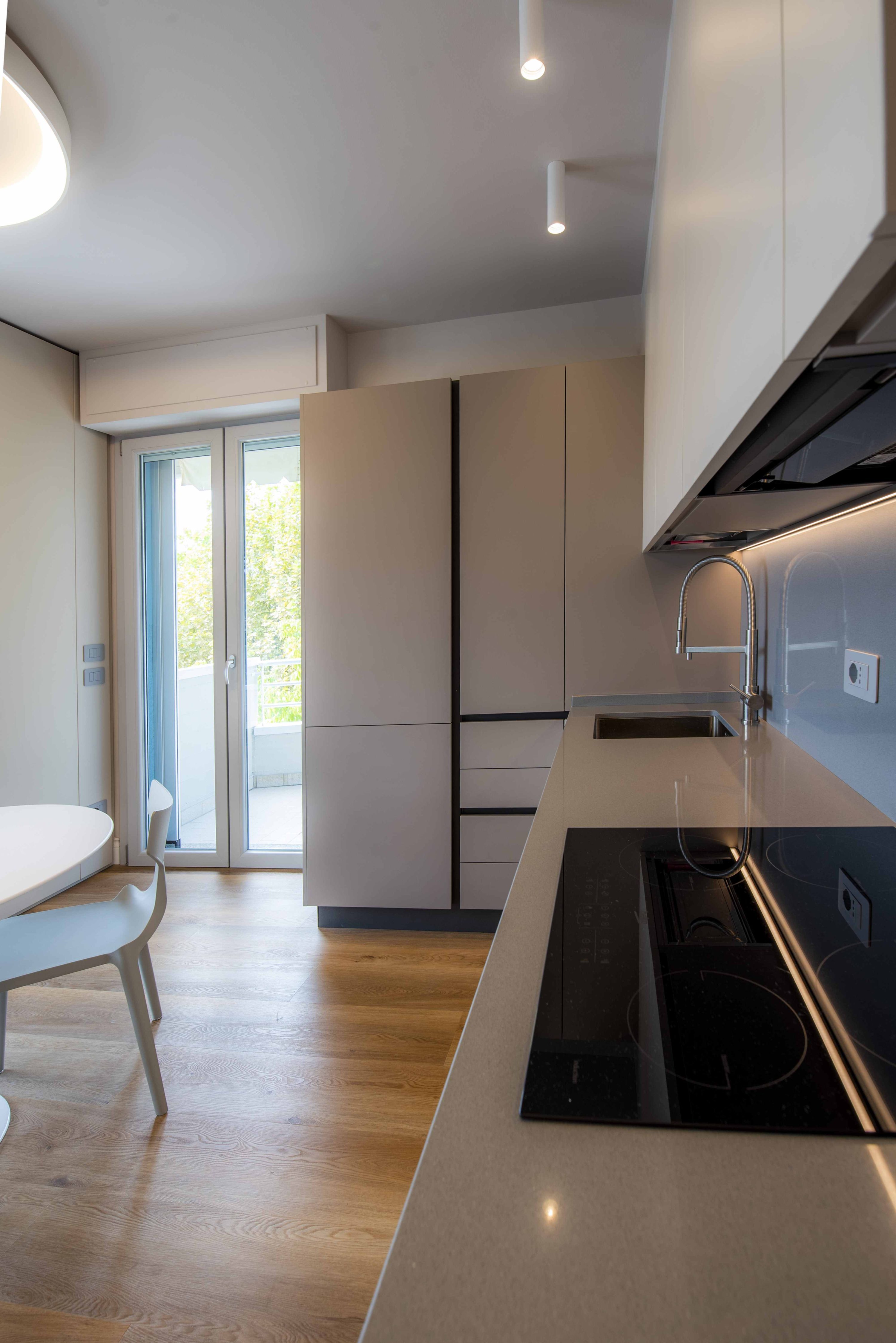cucina-moderna-minimalista-minimal-sabbia – MICHELE VOLPI STUDIO INTERIOR DESIGN Cucina moderna minimalista con piano a induzione nero, lavello sottotop e mobili color sabbia opaco. Vista su pavimento in parquet chiaro e porta finestra verso il balcone.