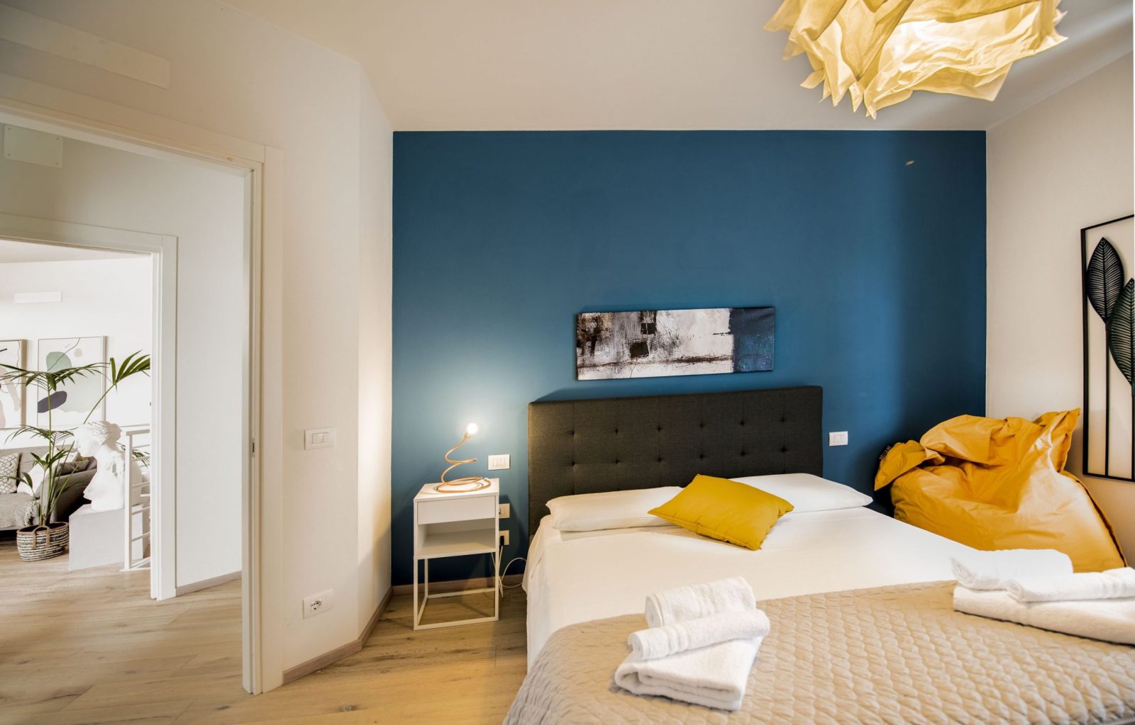 Camera da letto matrimoniale moderna con parete blu accesa, dettagli gialli, biancheria bianca e asciugamani puliti pronti per gli ospiti.