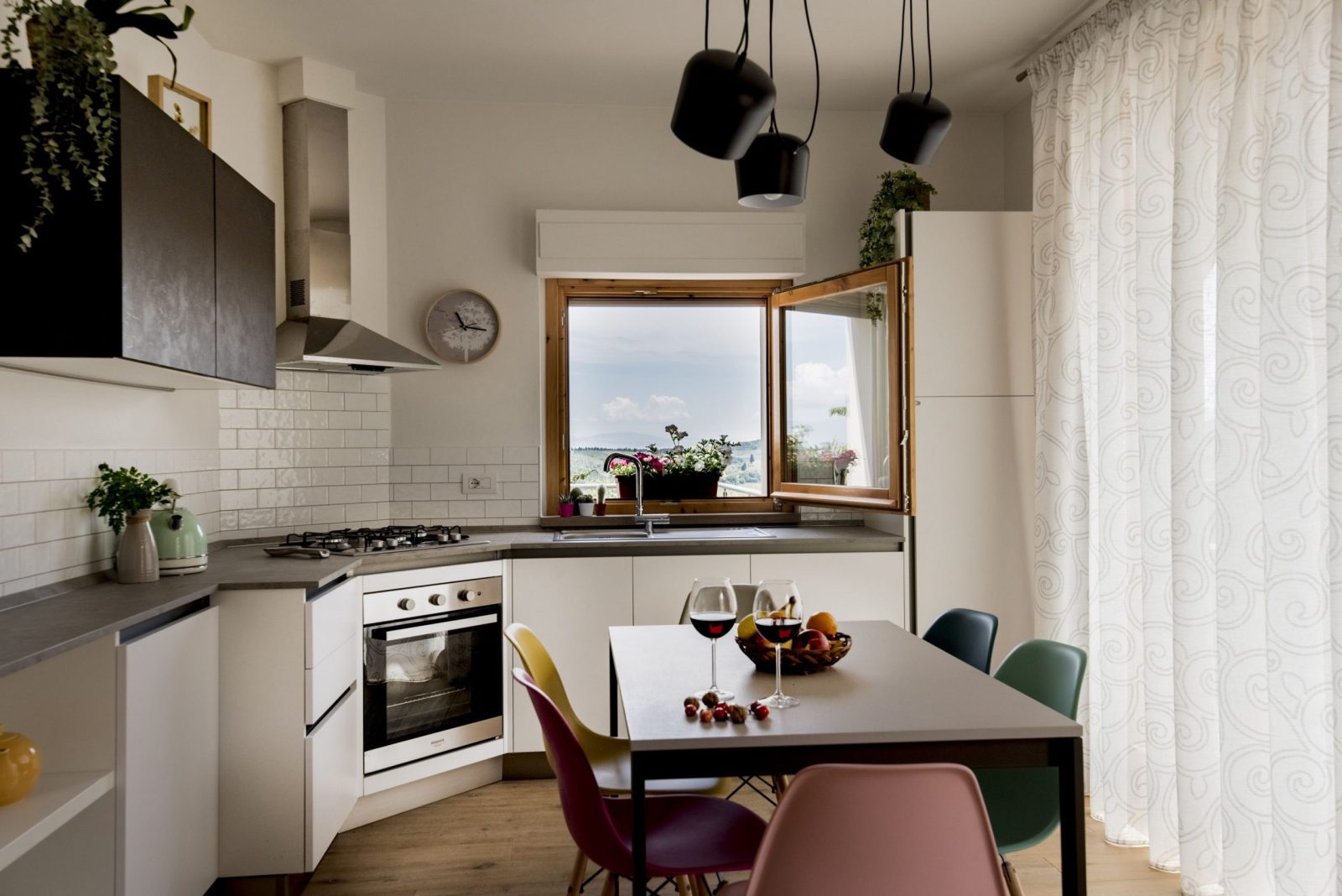 Cucina moderna minimalista con mobili bianchi, tavolo da pranzo con sedie colorate e finestra aperta con vista panoramica.