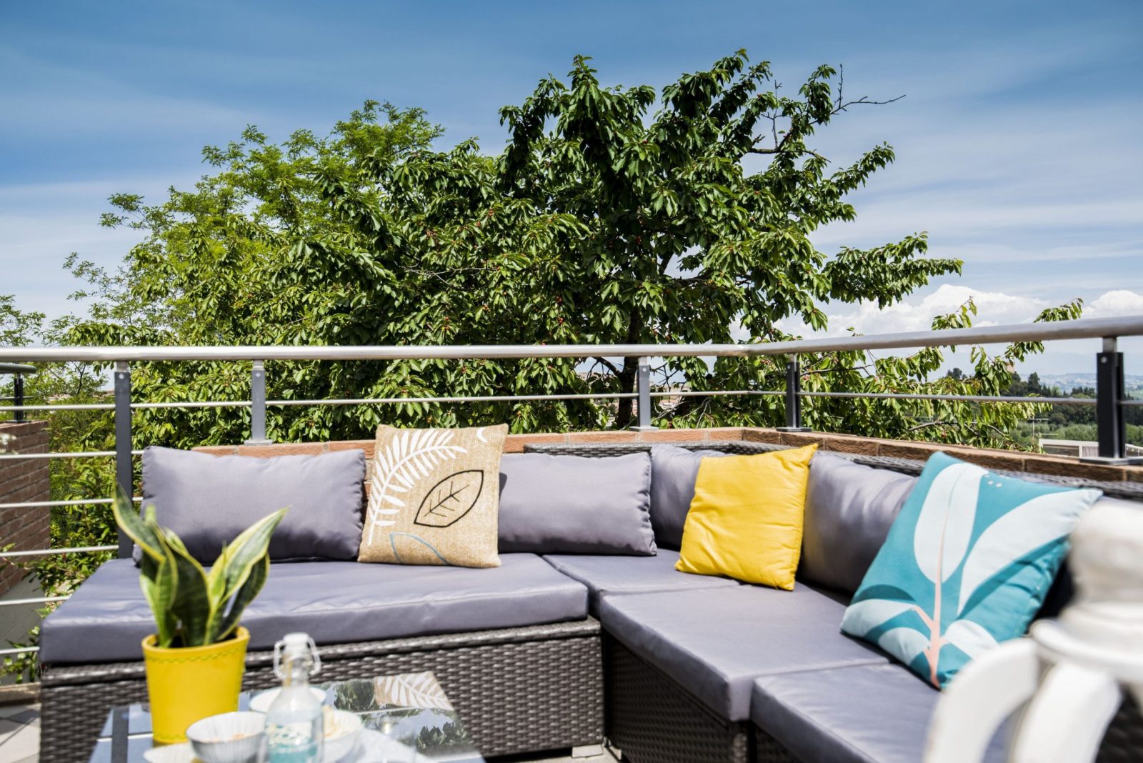Terrazzo esterno con divano angolare in rattan grigio, cuscini colorati e vista su un grande albero verde sotto un cielo azzurro.