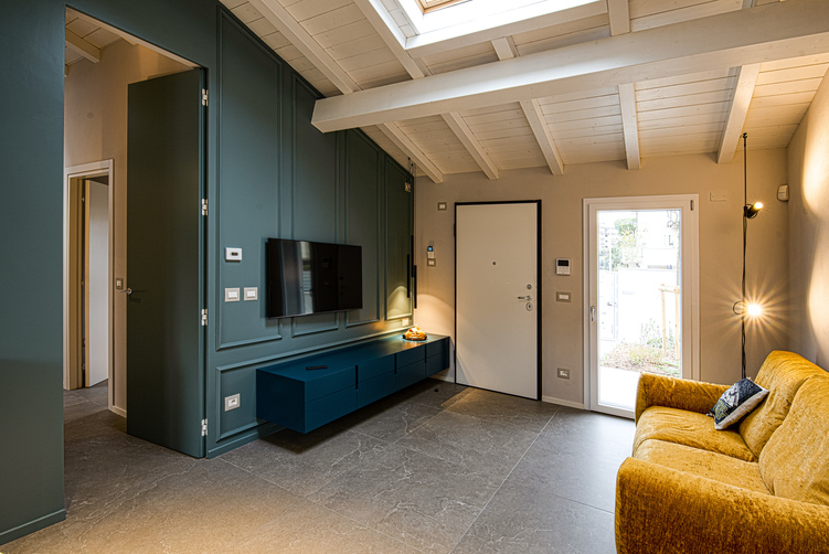 Soggiorno mansardato moderno con divano giallo senape, parete attrezzata blu petrolio a boiserie, soffitto con travi bianche a vista e pavimento in gres grigio scuro.